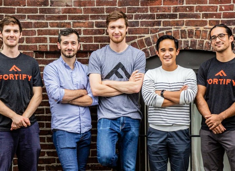 Five men in casual attire pose in front of a brick wall with their arms crossed.